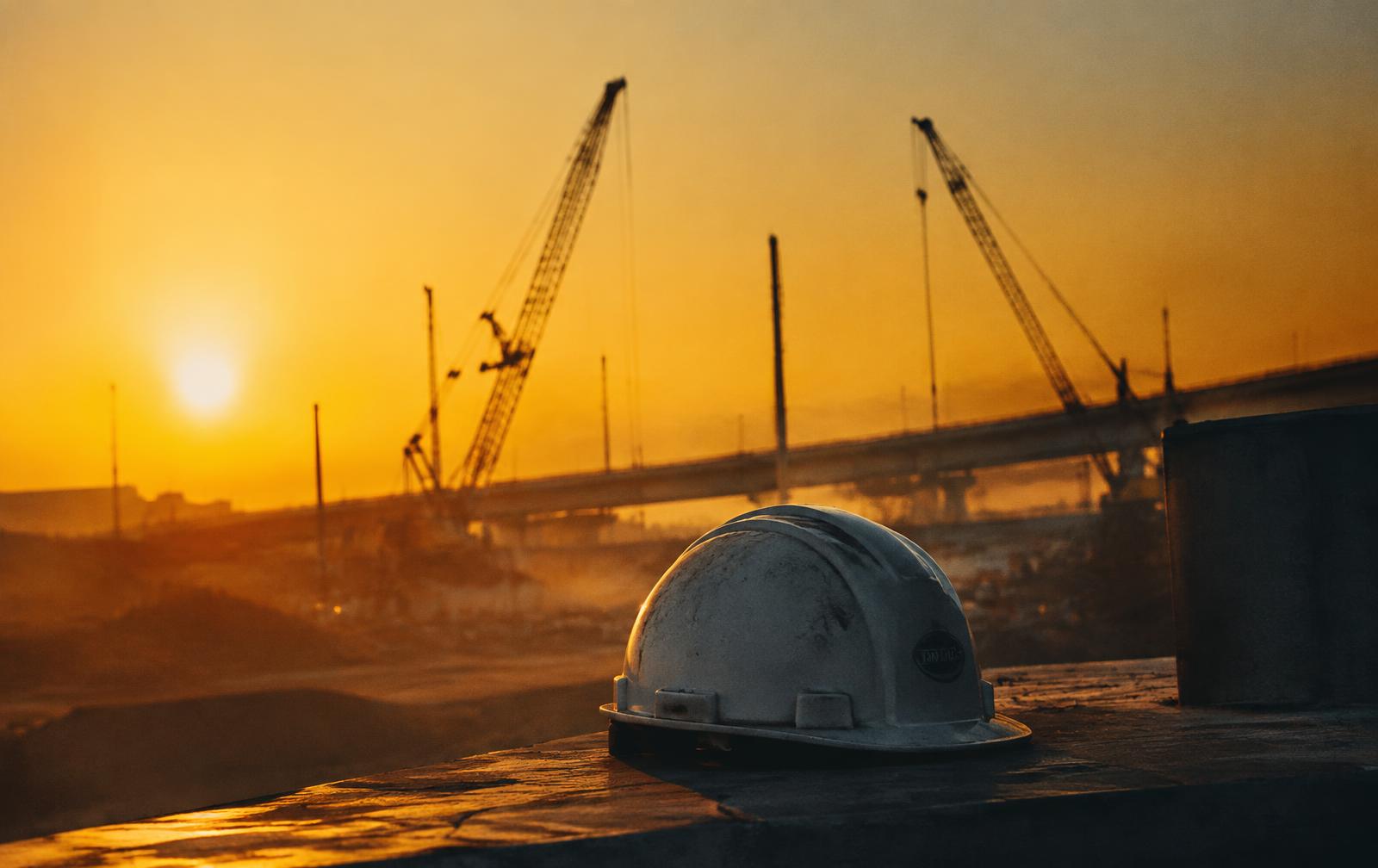 A white hard hat resting on concrete at a highway construction site at sunrise, cranes in the distance